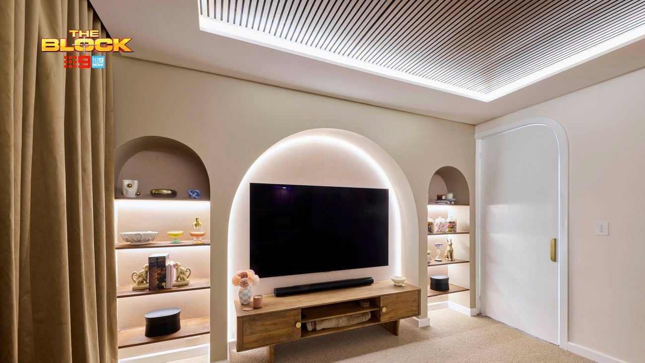 A modern living room with a large flat-screen TV mounted within a softly lit arched alcove. Below the TV is a wooden console table with decorative items and storage compartments. Built-in shelves in arched niches on either side of the TV display vases, books, and sculptures. The ceiling features recessed lighting and slatted paneling for a stylish finish.
