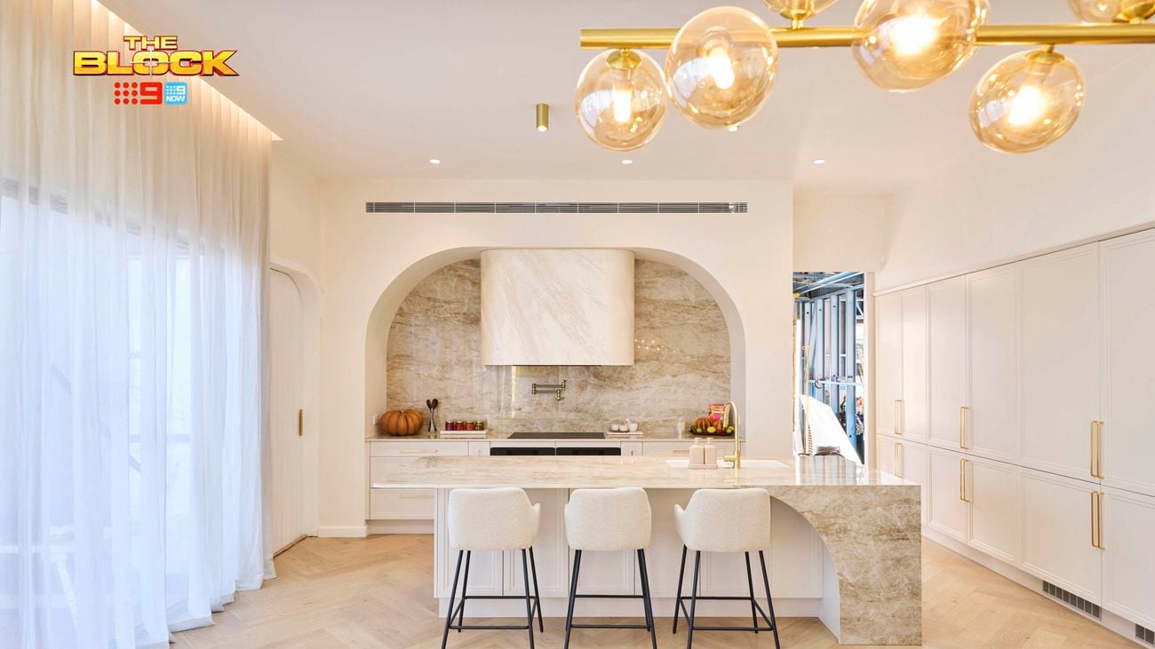 Bright, modern kitchen from The Block, featuring a marble island with four bar stools, arched range hood with stone backsplash, white cabinetry, gold fixtures, and a contemporary glass bubble chandelier.