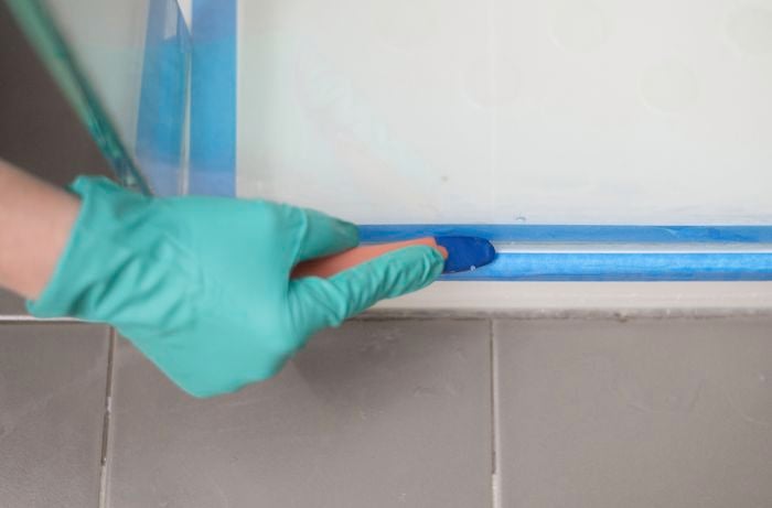 A hand in green gloves is using a scraper to smooth out freshly applied silicone.
