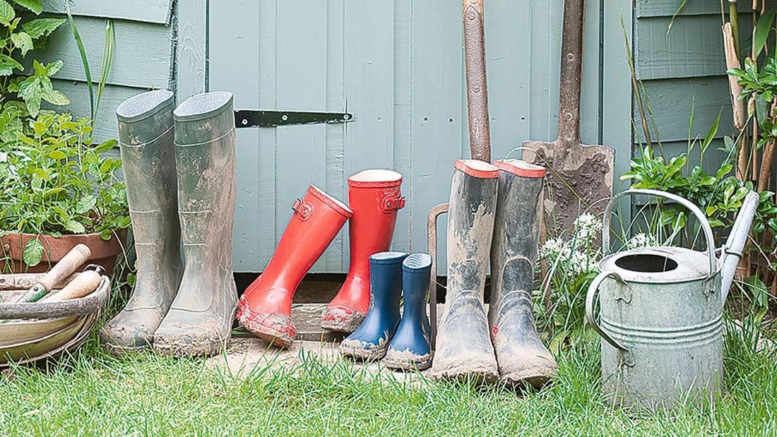 Gardening tools and equipment next to a shed, including gumboots, a shovel, a pitchfork and a watering can.