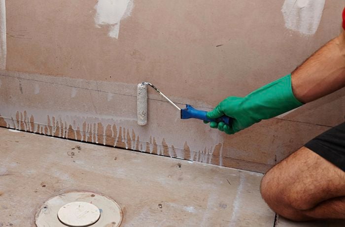 A person using a paint roller to apply primer along the bottom of a bathroom wall