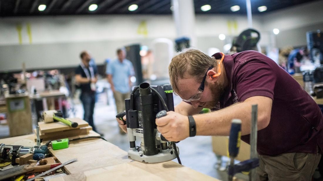 An apprentice wearing safety goggles in a workshop uses a plunge router on a piece of wood.
