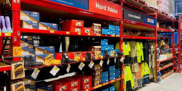 Hardware store workwear section featuring safety boots, high-visibility clothing, work apparel and accessories displayed on red industrial shelving.
