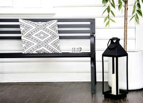 Front porch with a black bench and patterned cushion beside a tall potted tree. A large lantern with a candle on the floor, a wall light, and a striped bag hanging near the black front door.