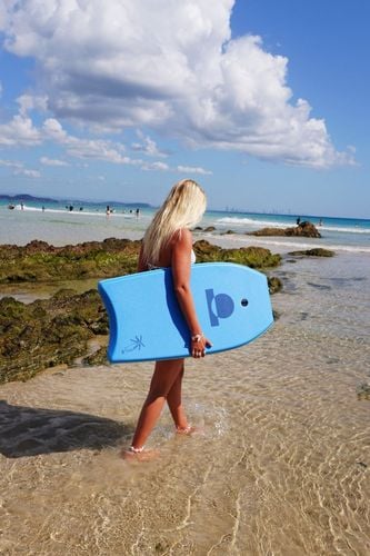 Woman carrying a blue Summer vibes bodyboard while walking through shallow water.