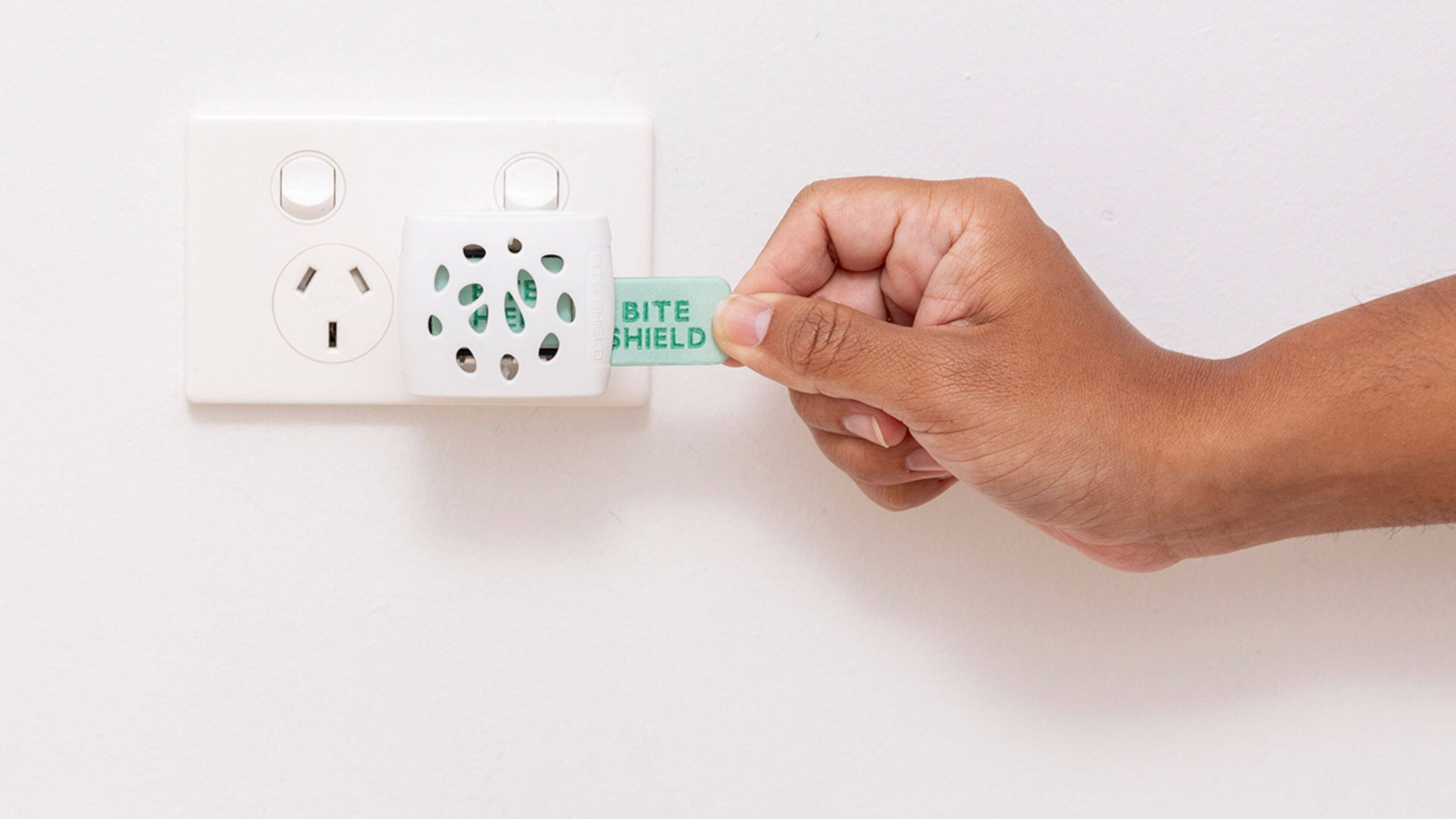 A hand holds the tag for Bite Shield plug in mosquito repellent plugged into a power point on a white wall.