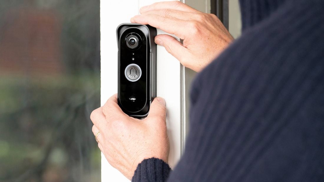 Close-up of a person installing a smart video doorbell on a white door frame, with the camera lens and doorbell button visible beside a glass panel.