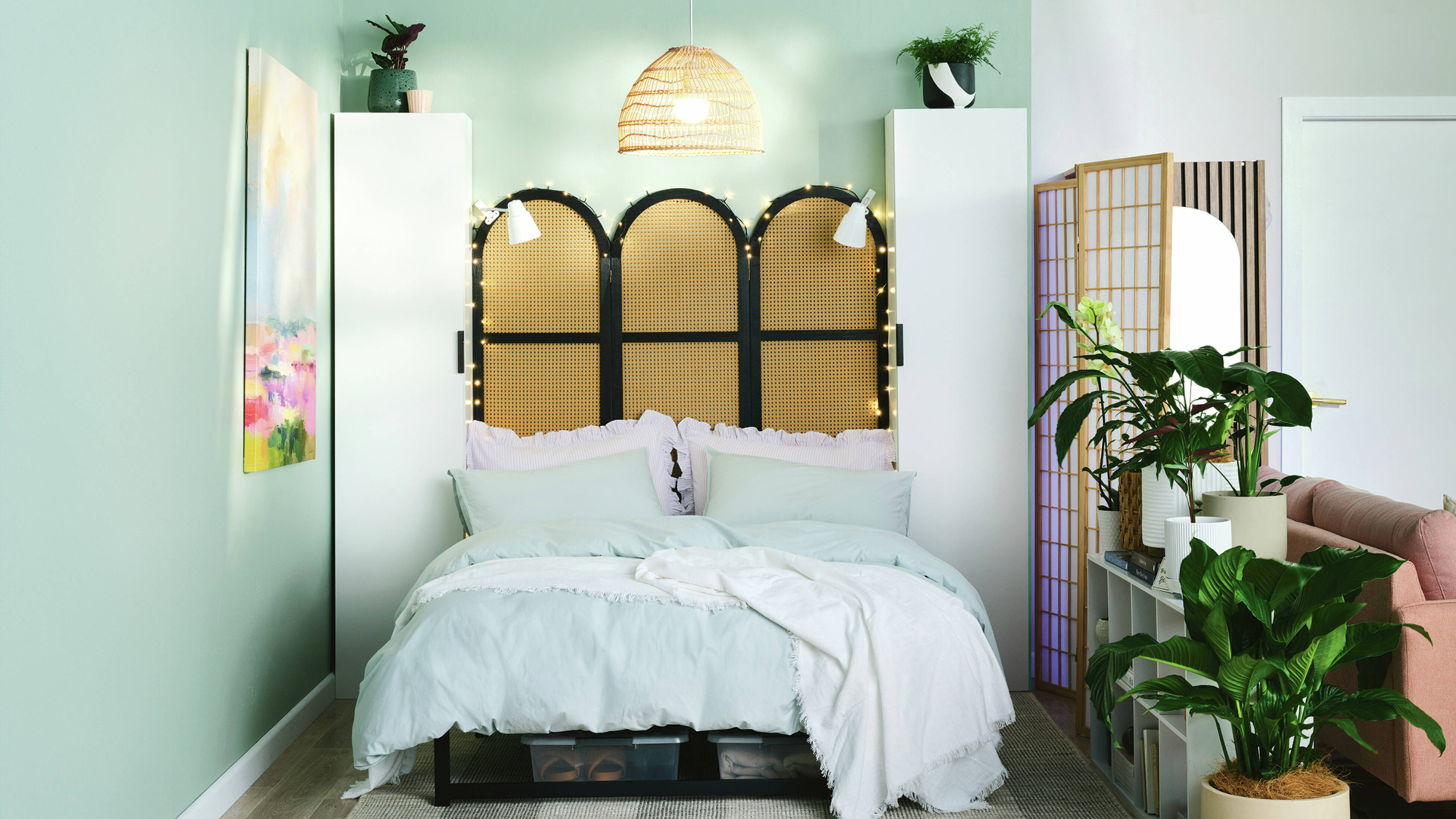 A bedroom with a bed with pale blue and white bed linen, a black screen as the bedhead, fairy-lights and two flatpack cabinets either side of the bed. Plus, plants in pots.
