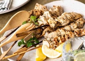 BBQ skewers on a white platter with a wooden serving fork and spoon, lemon wedges and a bowl of tzatziki on the side,