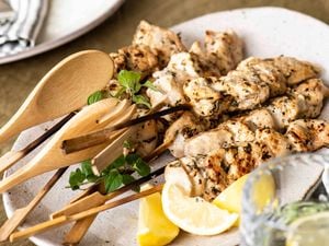 BBQ skewers on a white platter with a wooden serving fork and spoon, lemon wedges and a bowl of tzatziki on the side,