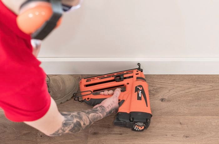 A person is using a nail gun to attach a skirting board to the wall.