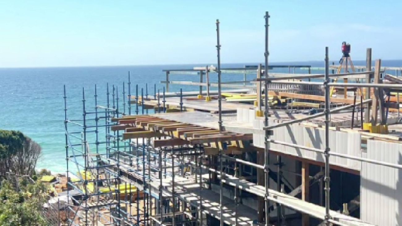 A building under construction with extensive scaffolding, situated on a cliff overlooking the ocean.