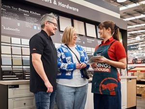Store team member in a Bunnings Warehouse apron shows kitchen cabinet samples to two customers standing beside a wall display of cabinet finishes at a Kaboodle kitchen planning station.