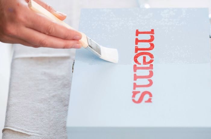 A person is applying a clear sealant with a brush over a painted wooden memory box to protect the design and give it a smooth, durable finish