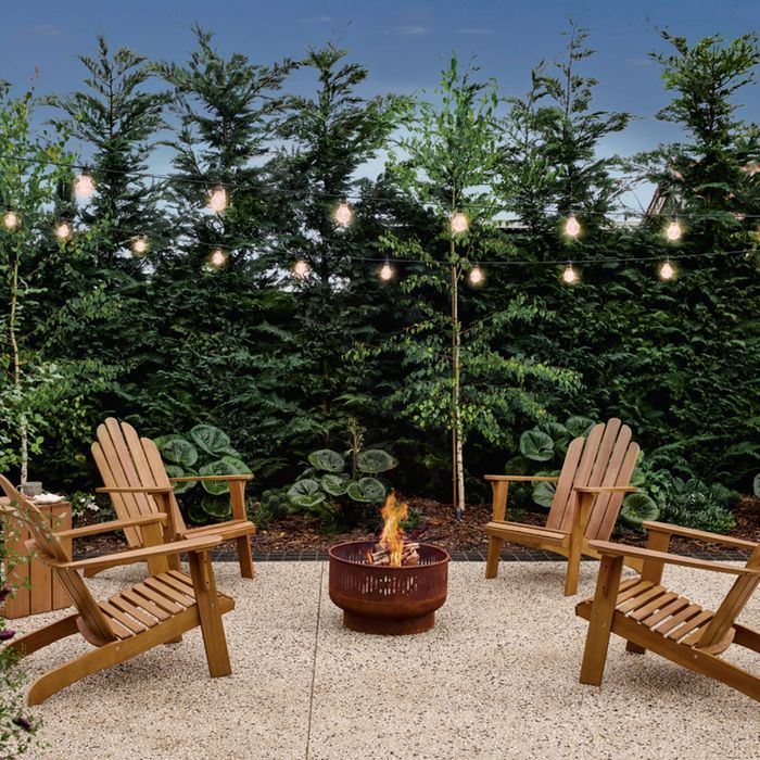 Outdoor patio seating area with four wooden Adirondack chairs arranged around a lit metal fire pit on a paved surface. Warm string lights hang overhead, with dense evergreen hedges and leafy plants creating a lush garden backdrop at dusk.