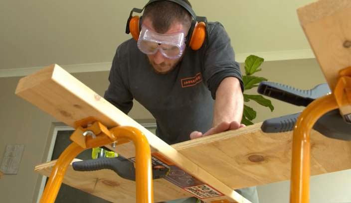 A man drilling a screw into a piece of timber being held by a saw horse