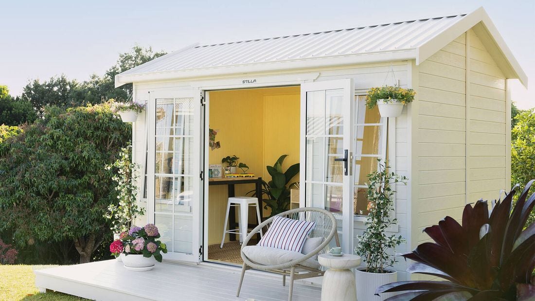 An angled view of a large timber shed painted white with a front deck featuring hanging baskets, pot plants and a chair sits in a garden setting. The open French doors show the yellow interior, a desk and stool.