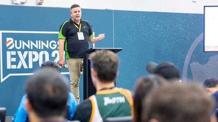 A presenter stands at a podium addressing an audience at a Bunnings Expo seminar, with attendees seated and looking toward the stage and display screens.