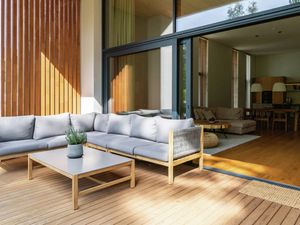 Modern outdoor wooden deck with a grey sectional sofa and coffee table, looking into a bright interior space through large sliding glass doors.