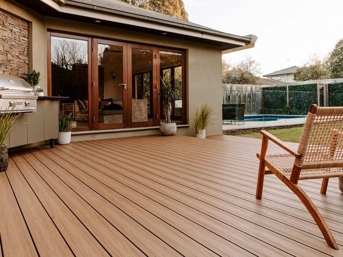 The back of a house with decking that has a barbecue, plants and a cane chair on it.