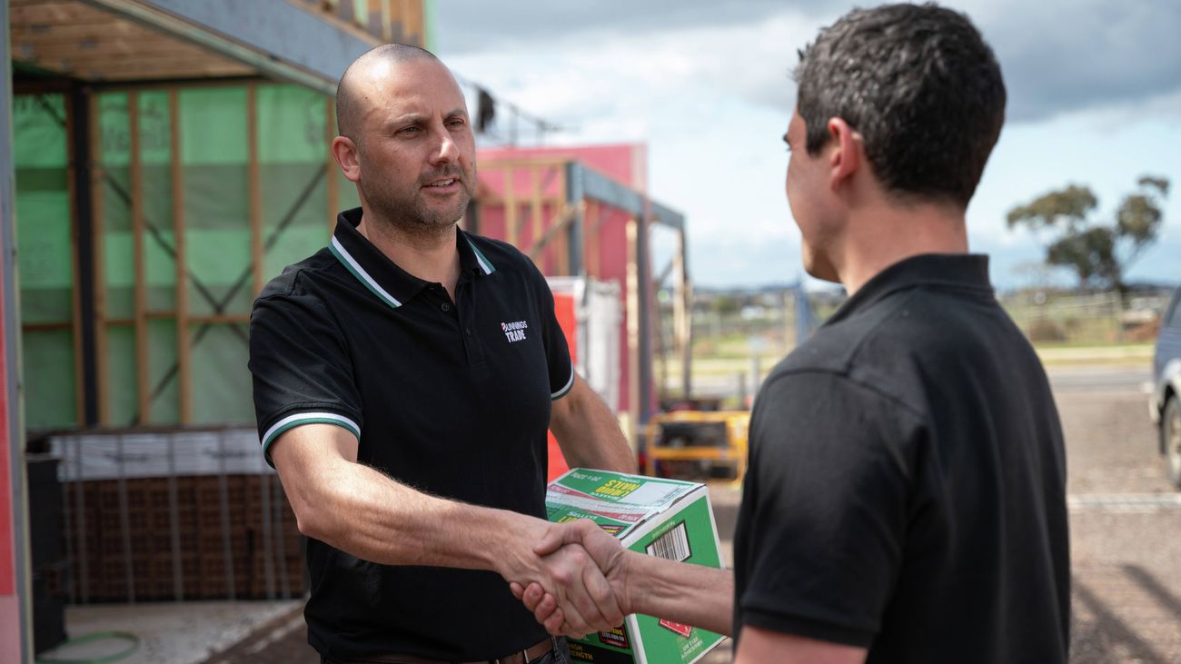 An image of two men in black t-shirts taken from the waist up. One is facing front-on, the other side-on, as they stand on a building site shaking hands