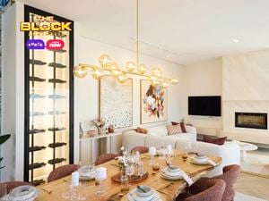 A modern living and dining area featuring a dining table set with plates, glasses, and candles, surrounded by six chairs. A contemporary chandelier with spherical lights hangs above. In the background, a white sofa with colorful cushions sits beneath two large abstract artworks. A fireplace with a TV above it is on the right, and a tall wine rack filled with bottles is on the left.