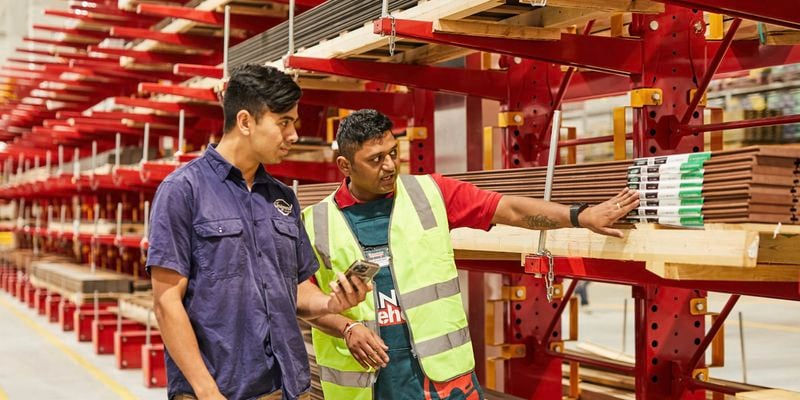 Inside bunnings warehouse, two team members stand beside red industrial shelving stacked with timber lengths. One, wearing a hi‑vis safety vest, gestures to product labels on the rack. The other, in a work shirt, holds a mobile device and reviews details as they discuss stock and product options.