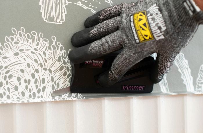 A gloved hand is using a wallpaper trimmer to trim the excess paper at the bottom of the wall