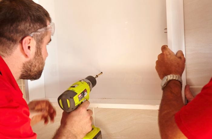 One person driving a screw through a cabinet while another person holds it against a tiled wall