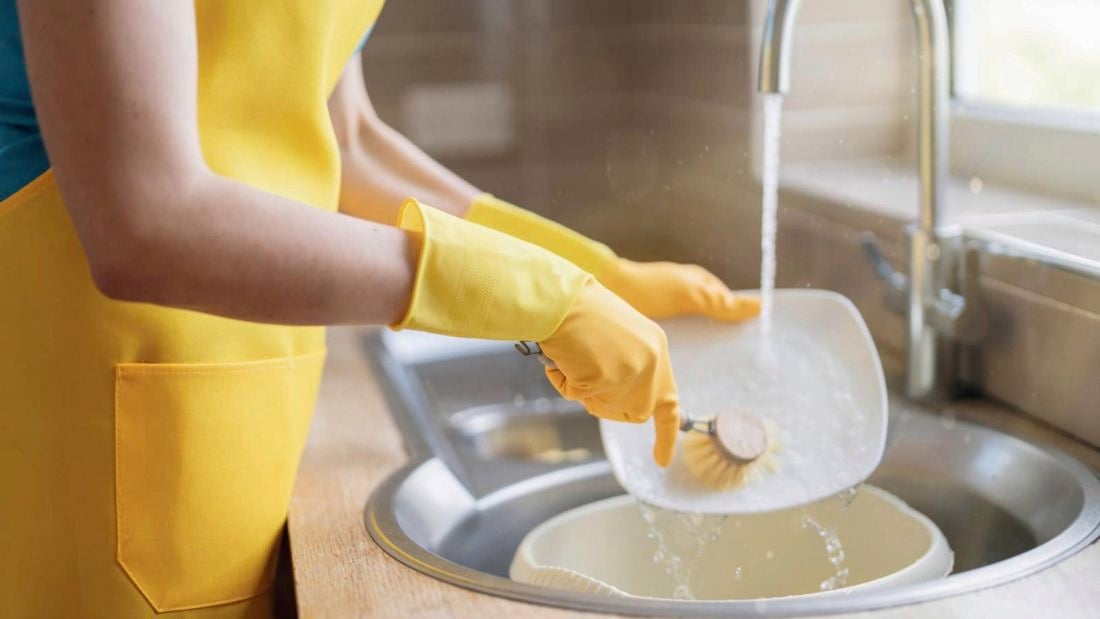 A woman wearing yellow dishwashing gloves and a yellow apron uses a scrubbing brush to wash dishes in a sink