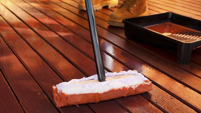 Applying timber deck stain with a deck applicator pad on a wooden outdoor deck. A paint tray sits nearby while a person wearing work boots stands in the background, showing the process of staining and protecting outdoor timber decking.
