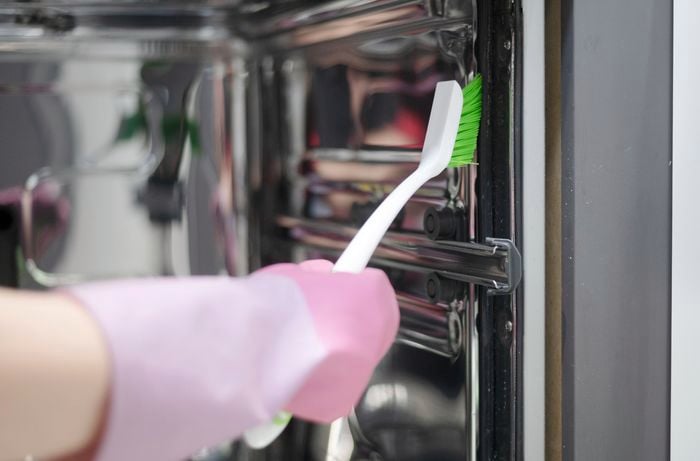 A hand with pink rubber gloves is using an old toothbrush to remove grease residue from the edges of the dishwasher.