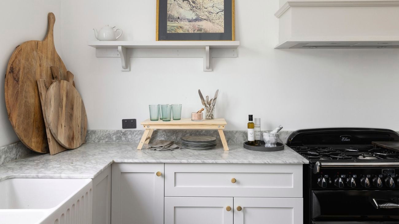 A white kitchen with marble benchtops, featuring a gas oven, chopping boards, and a timber picnic tray table with kitchenware.