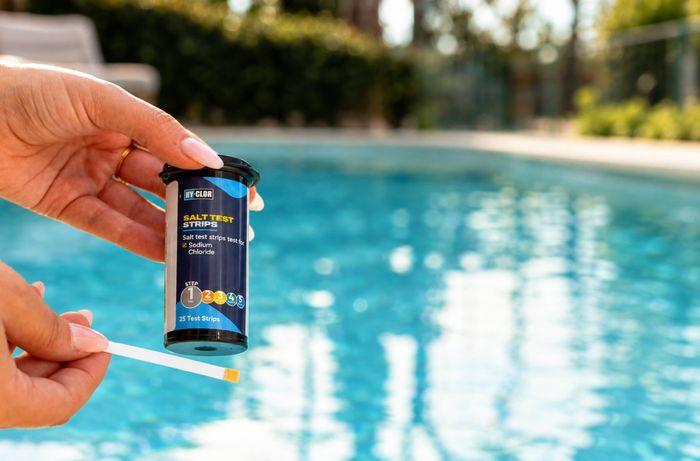 Close-up of a person holding a pool water testing strip and a container of salt test strips beside a swimming pool.