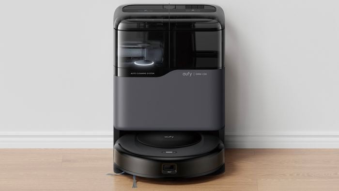 Eufy Omni C20 robot vacuum docked in a self-cleaning station with a dark gray and black finish, positioned against a light gray wall on a light wood floor. The compact docking unit features enclosed water tanks and automatic cleaning components, with the round robot vacuum resting at the base.