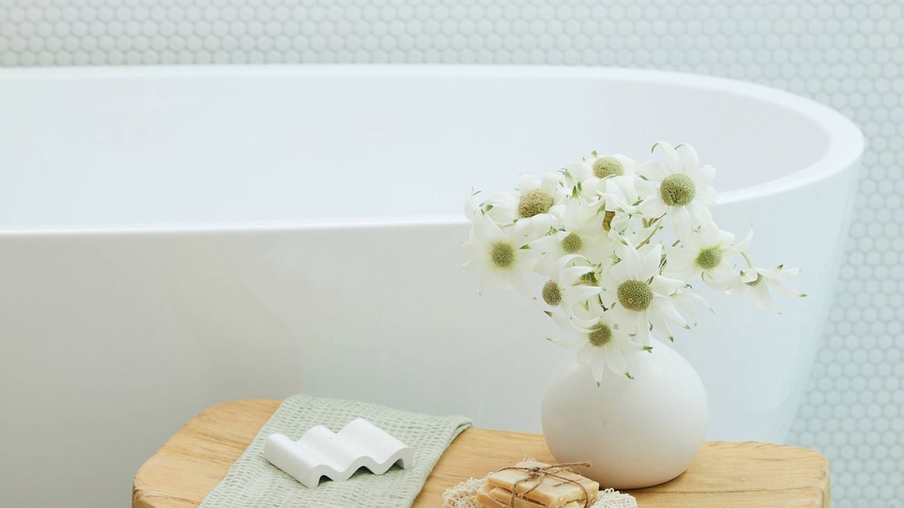 Bathroom with a white freestanding bathtub against a white hex-tile wall and window. A small wooden stool holds a light green towel, white zigzag soap, round white vase of white flowers, and a loofah.
