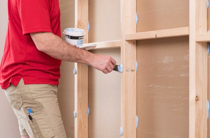 A person in a red T-shirt applies stud adhesive to wall studs with a spatula.