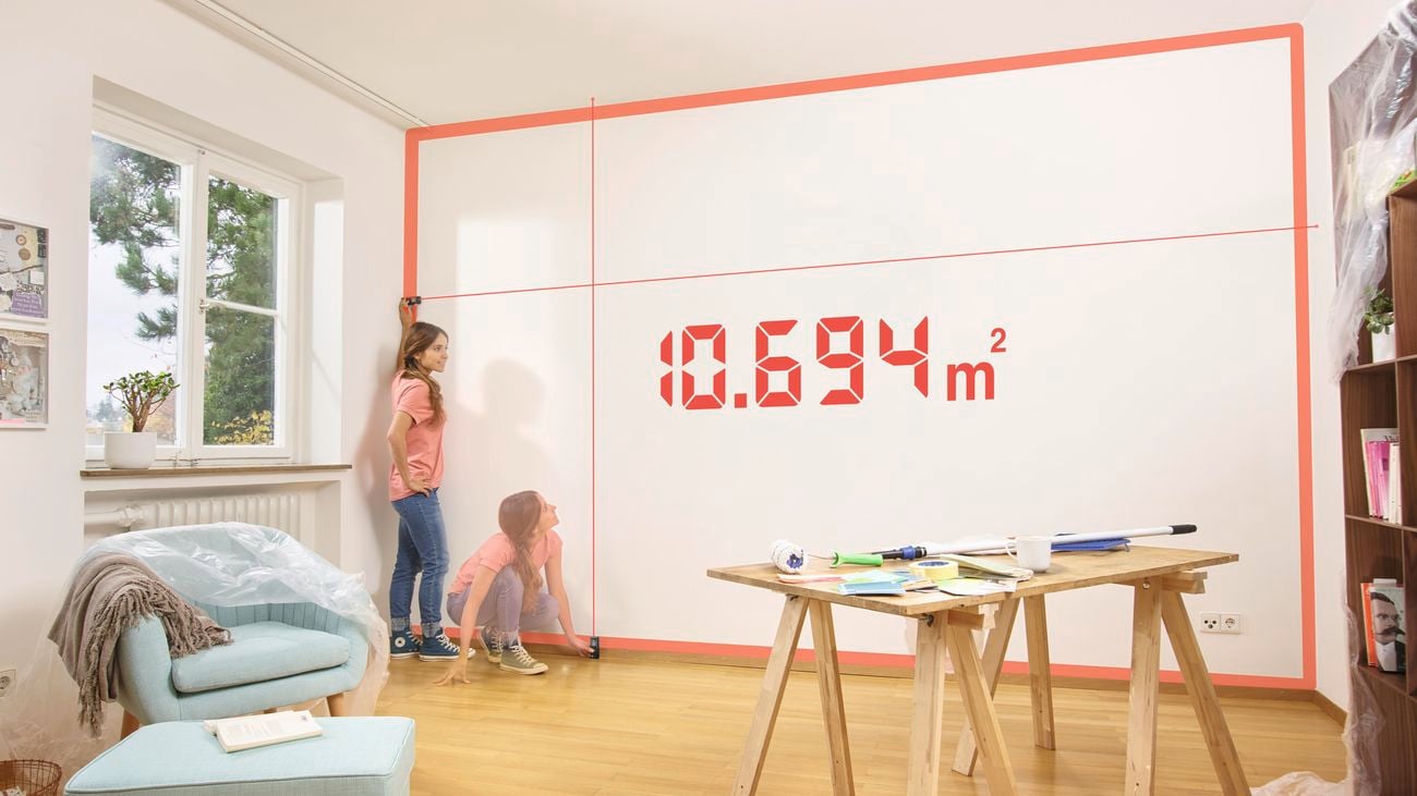Living room with a chair, footstool, bookcase and table, with a person using a Bosch Zamo Laser Measure Set to measure the size of a blank wall.