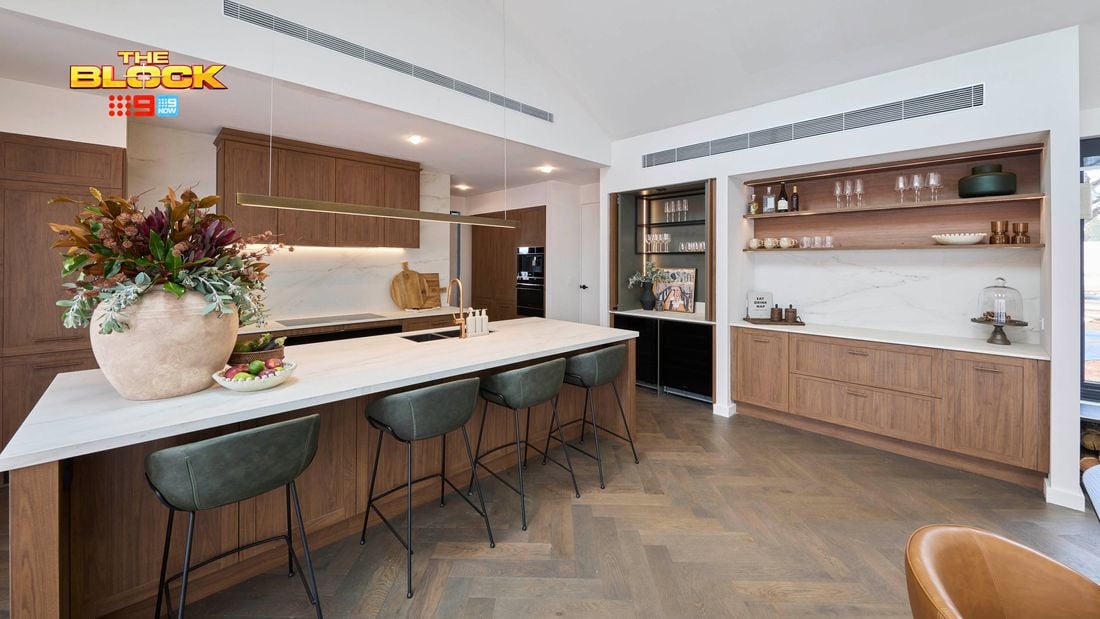A modern white kitchen with brown cupboards, bar stools at the benchtop with a vase with flowers in it and two bowls with items in them on it.