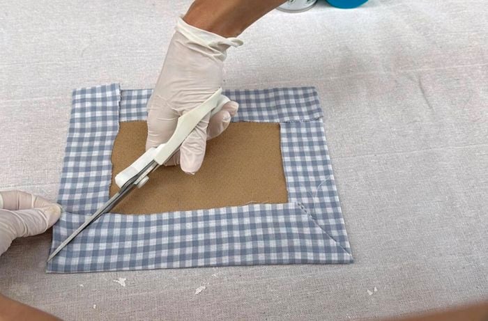 Hands wearing gloves, cutting a square piece of cardboard covered in blue and white checkered fabric