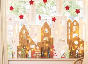 A Christmas window display featuring a homemade cardboard Advent calendar village on the sill, framed by waterfall lights and red craft stars.]