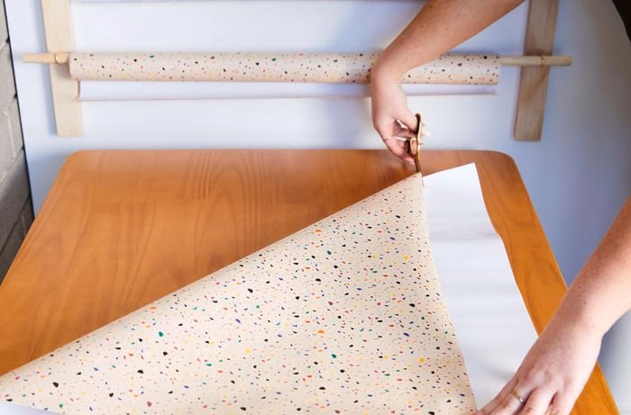 A pair of hands is cutting a folded sheet of wrapping paper with scissors on top of a wooden table.