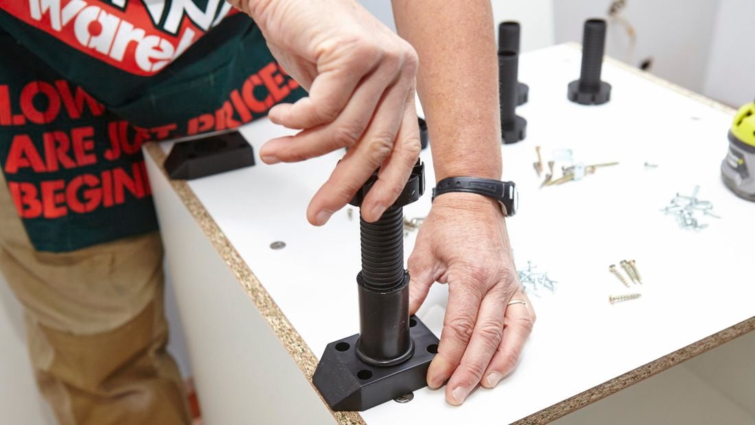 A person screwing a leg into a base plate on the bottom of a cabinet