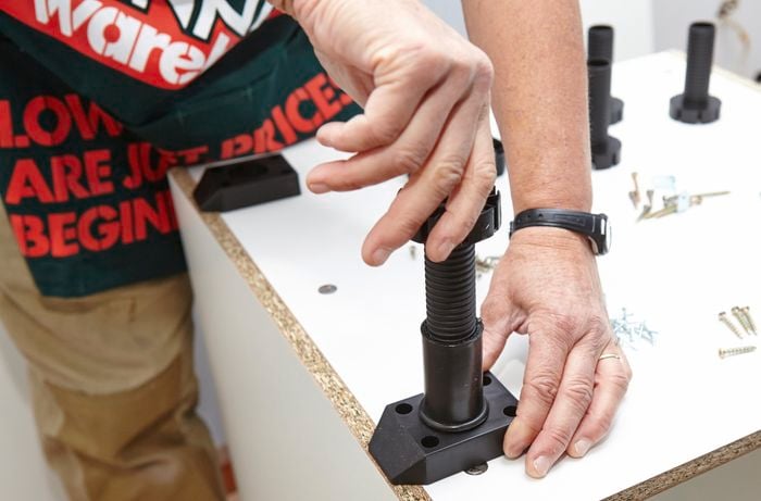 A person screwing a leg into a base plate on the bottom of a cabinet