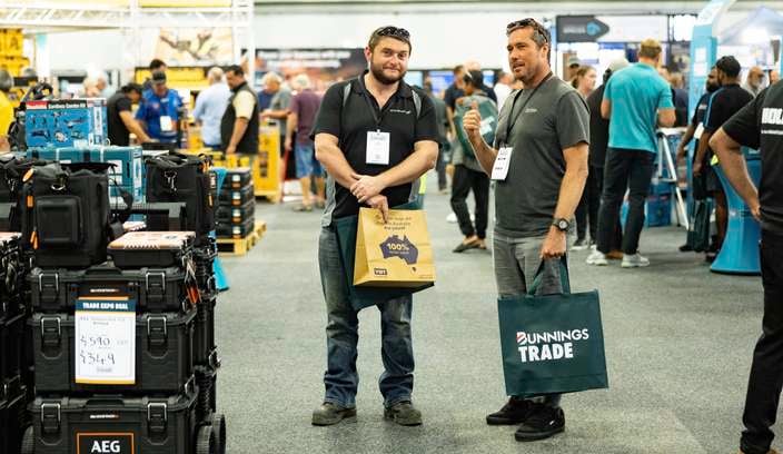 Two people  at the Bunnings Trade Expo stand among tool displays, holding branded shopping bags, with DeWalt signage and other exhibitors visible in the background.