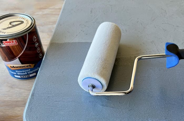 A hand using a paint roller to apply varnish onto a painted door panel, a tin of varnish next to it.