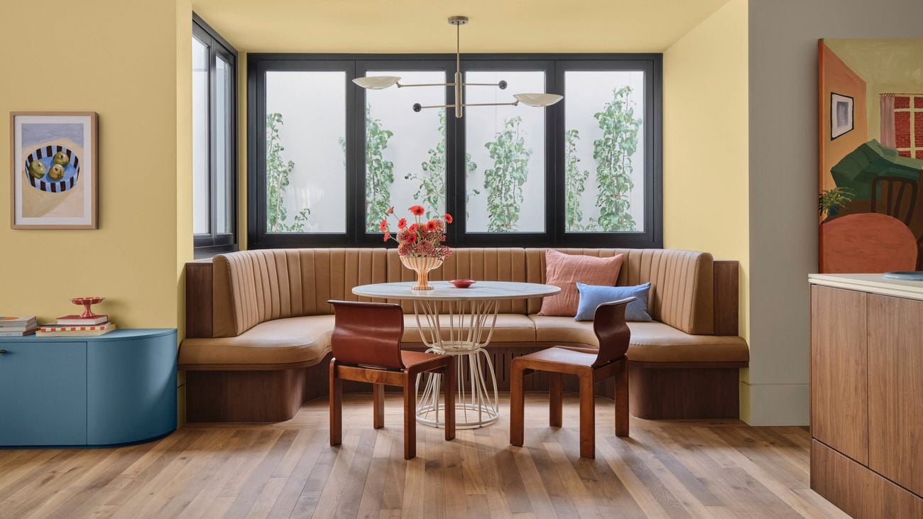A stylised eating area set in a bay window with circular tan seating, a table with two timber chairs, wall paint in Dulux Germania and other colourful furnishings.