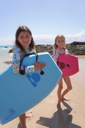 Two smiling girls holding blue and pink bodyboards on the beach.