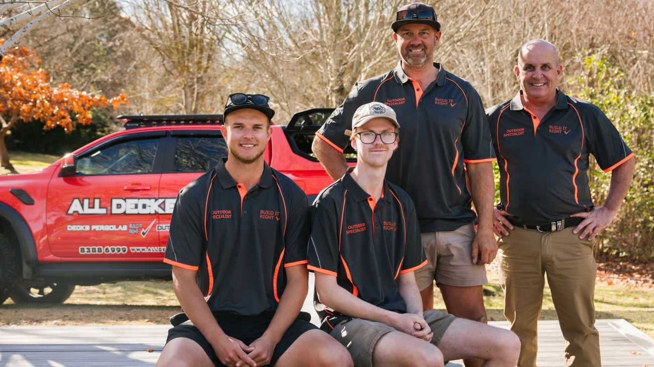 Builder Glen Campbell and four tradesmen in his team sit on a bench and a composite deck, a red ute and trees are behind them