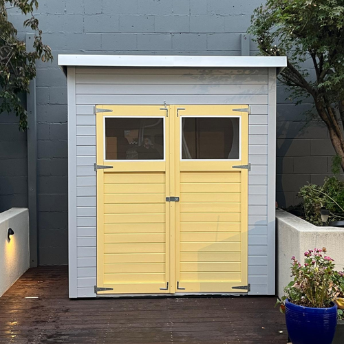 Compact grey storage shed with a skillion roof and pale yellow double doors featuring upper windows, situated on a timber deck.